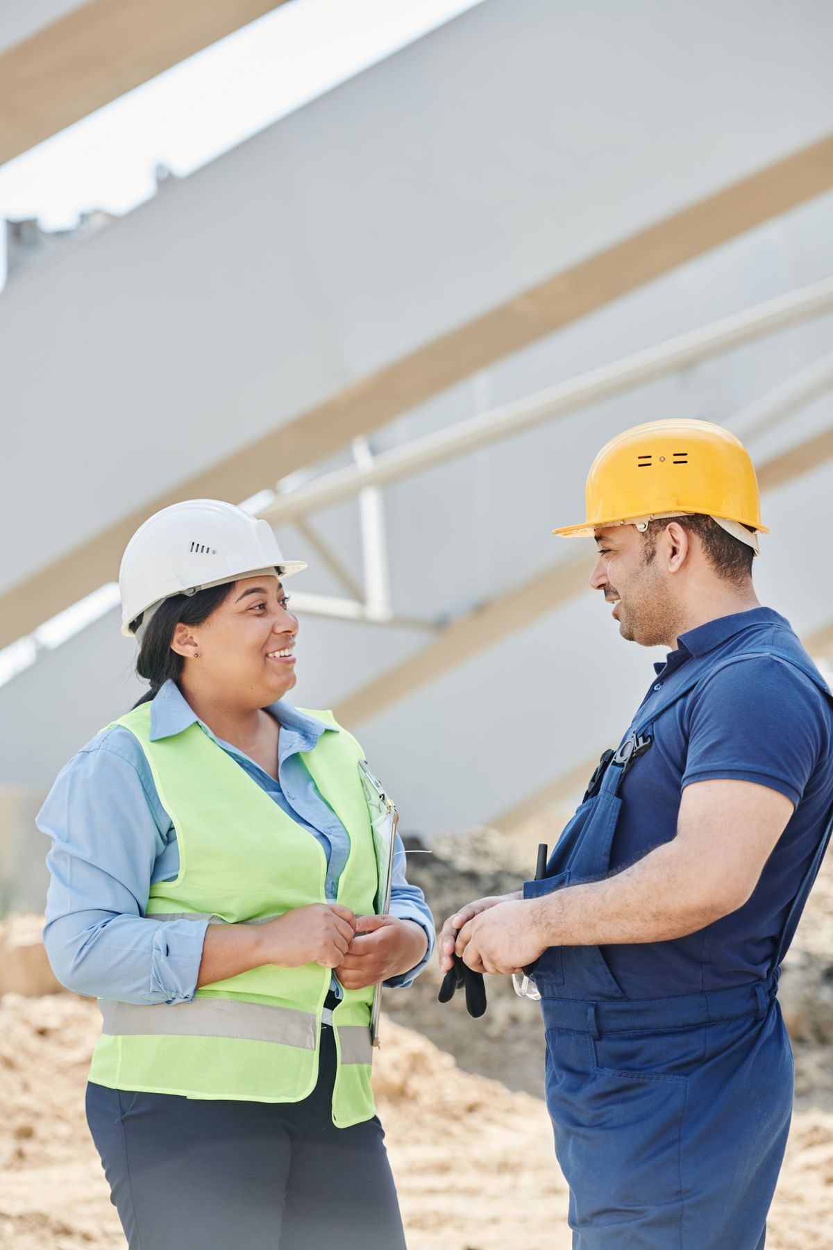 Engenheiro e engenheira conversando e sorrindo, com capacetes de segurança, em uma obra.